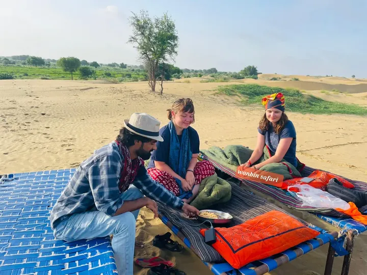A guide serving food to two guests sitting on charpoys on the open sand at Siala
