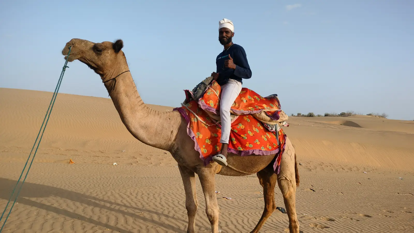 A cameleer in a white turban riding his camel across an open dune