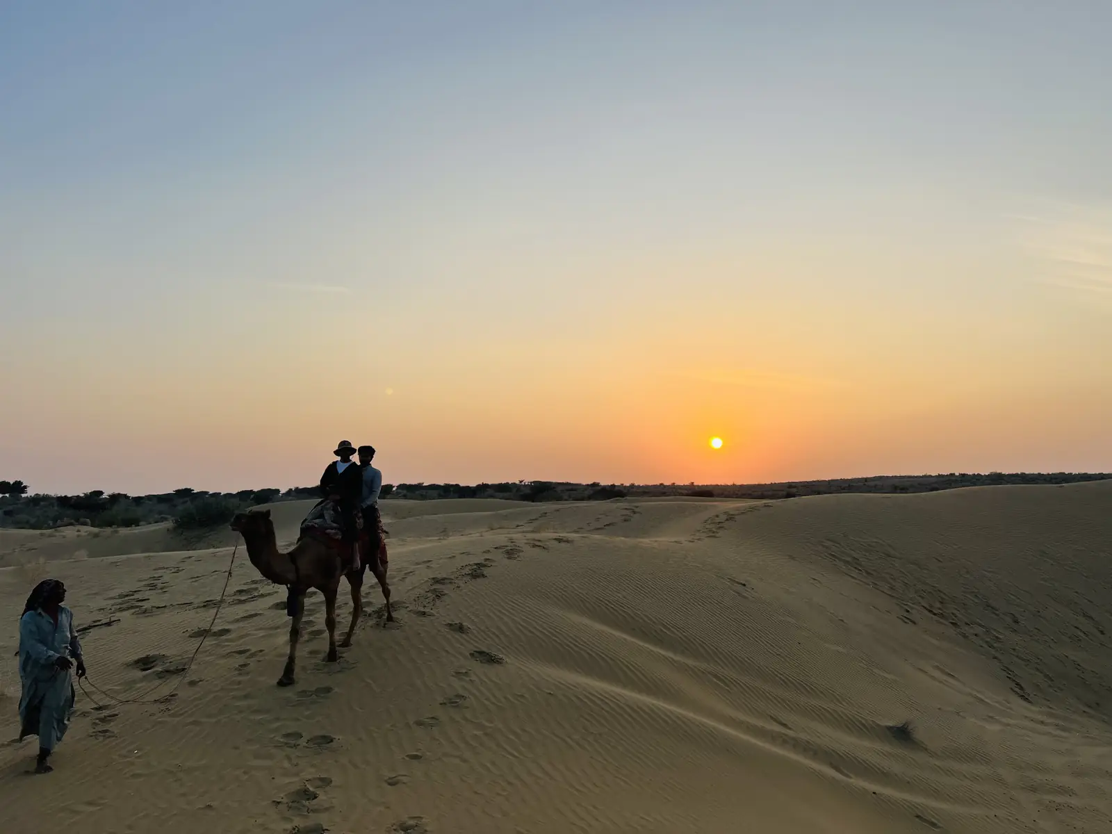 A camel and its rider with the camel man walking alongside as the sun goes down