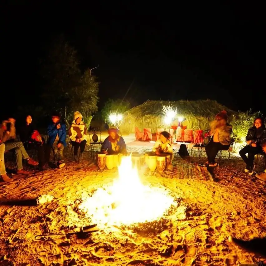 Guests sitting around the campfire while two musicians play, the thatched shelter lit behind them