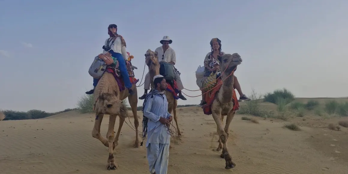 Three guests riding camels led on foot by a cameleer across the dunes